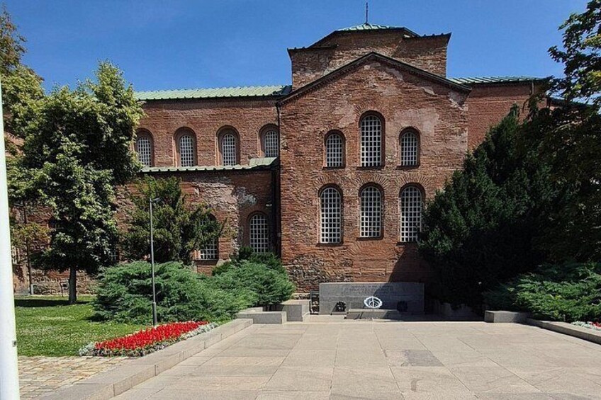 A large brick building with arched windows and a central dome, surrounded by a paved courtyard and greenery. 2