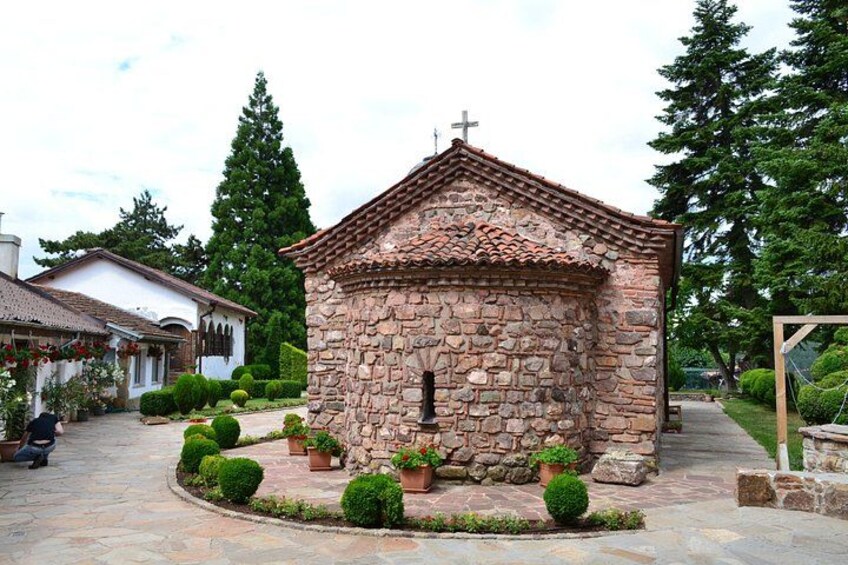 A small stone chapel with a cross on top, surrounded by a stone pathway and trimmed bushes. 3