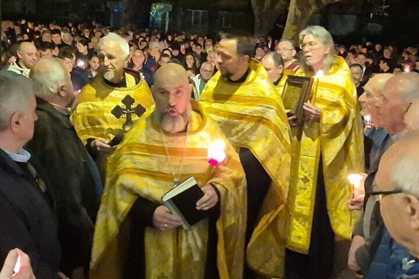A group of people in yellow robes holding candles and books, gathered in a nighttime outdoor setting. 5