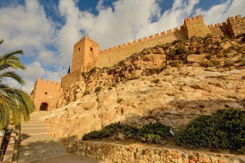 A historic stone fortress with a tower, a staircase leading up to the entrance, and a palm tree in the foreground. 2