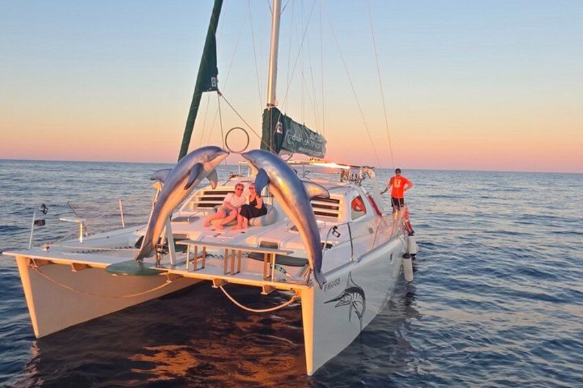 A sailboat with a covered deck, two people sitting, and a person standing on the stern. 5