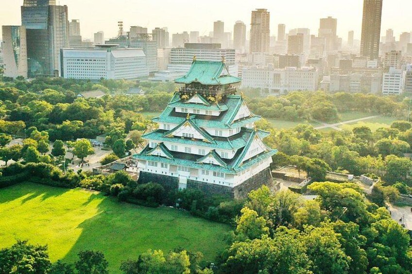 Ett traditionellt japanskt slott med flera våningar, omgivet av frodig grönska och med en stadsbild i bakgrunden. 1