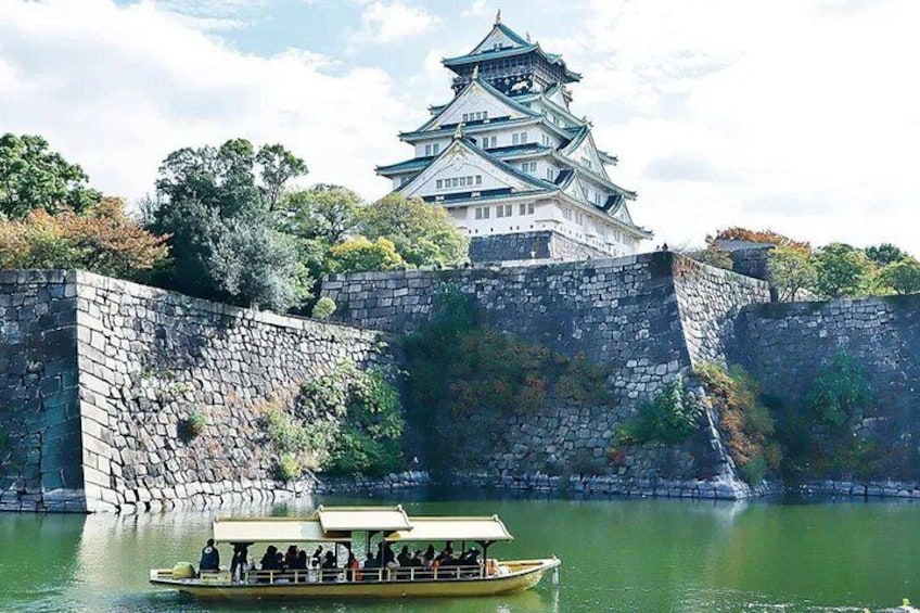 A castle with multiple tiers, a stone wall, and a boat with people on a calm body of water. 5