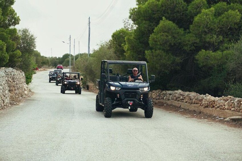 A convoy of off-road vehicles driving on a rural road surrounded by trees and stone walls. 3