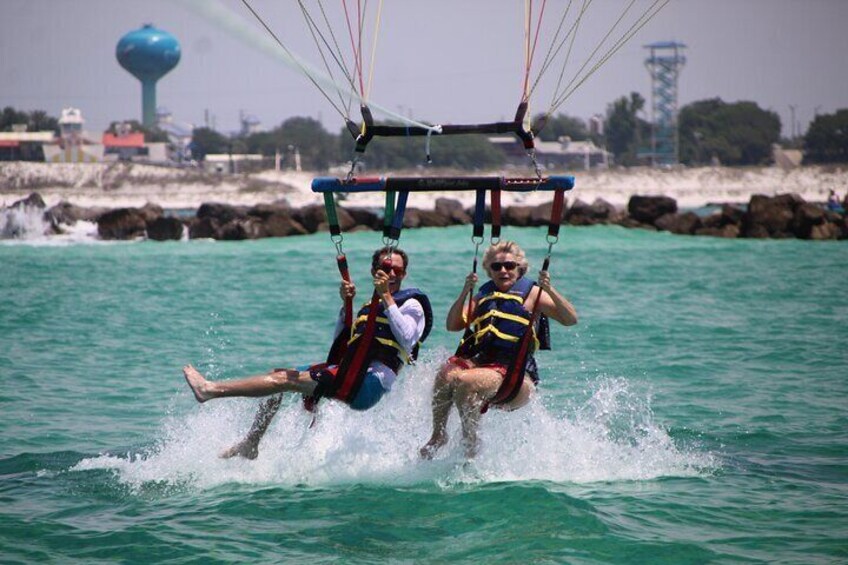 Two people parasailing over turquoise water with a rocky shoreline and a water tower in the background. 4