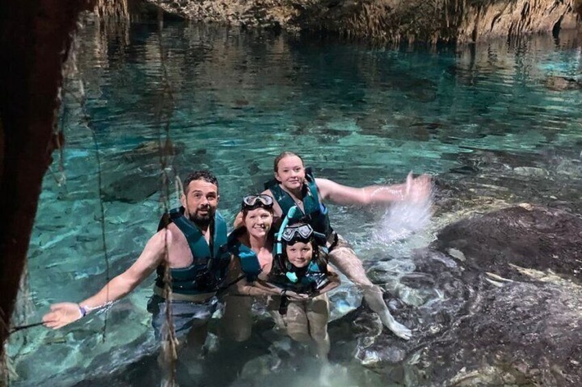 Three people in clear water, wearing life jackets and goggles, with a rocky cave background. 4