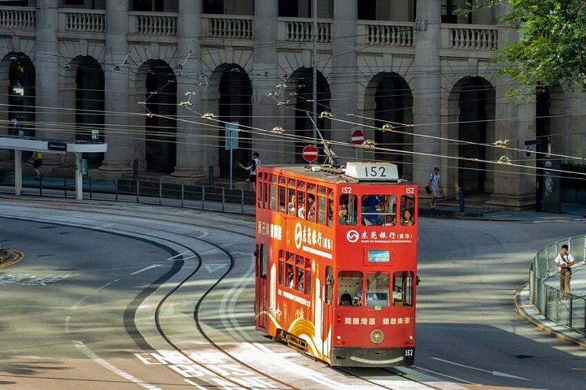 Eine rote, zweistöckige Straßenbahn mit der Nummer 152 fährt auf einer gewundenen Straße vor einem großen Gebäude mit Rundbogenfenstern. 3