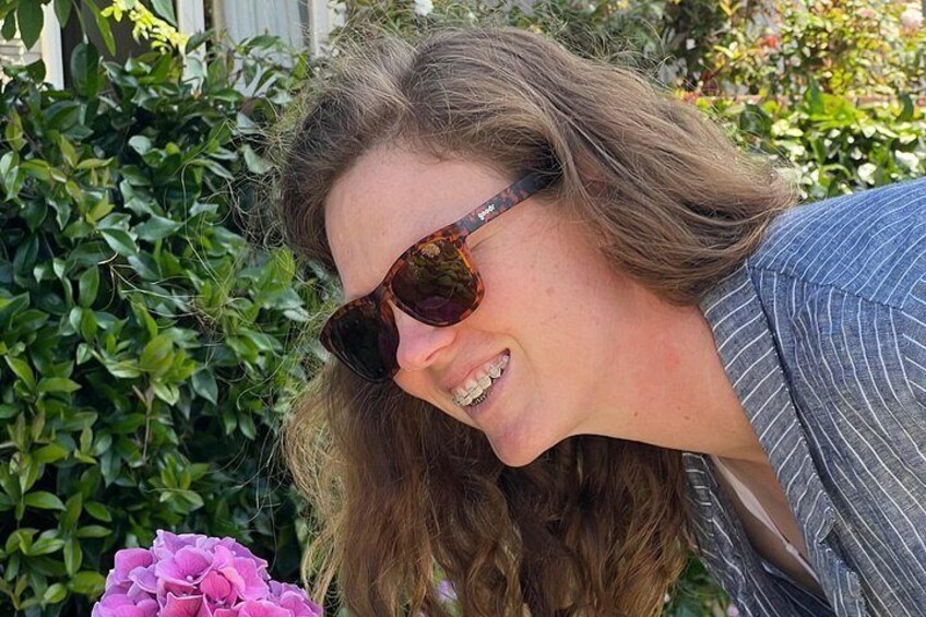 A person with long hair wearing sunglasses and a striped shirt, standing in front of a green hedge with pink flowers. 4