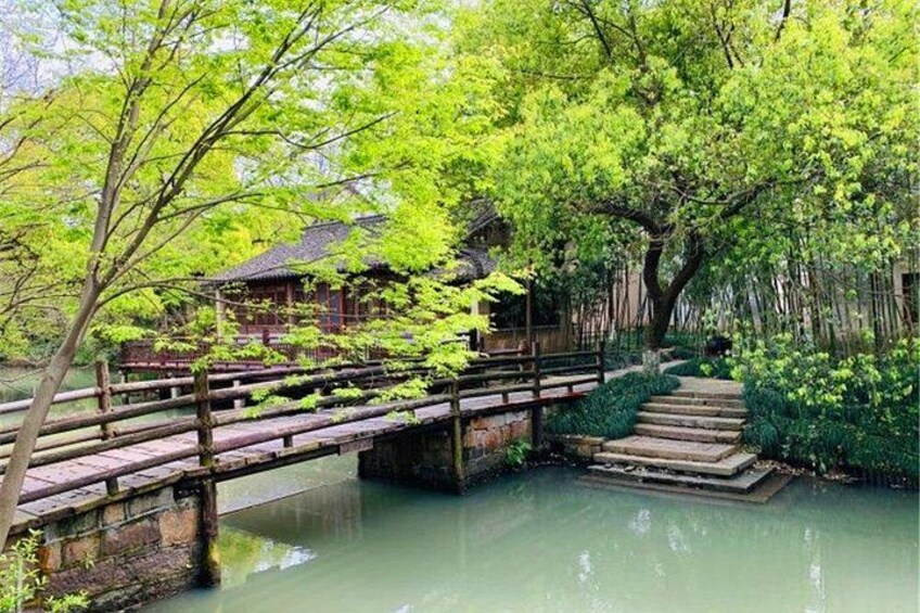 Un puente de madera tradicional cruza un cuerpo de agua tranquilo, rodeado de abundante vegetación y un edificio tradicional. 2
