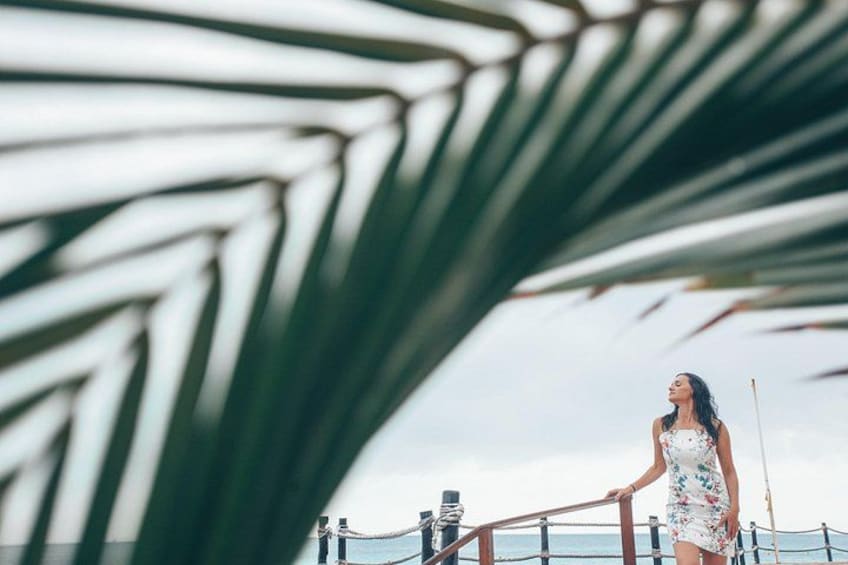 A person standing on a pier with a railing, palm leaves in the foreground, and a body of water in the background. 2