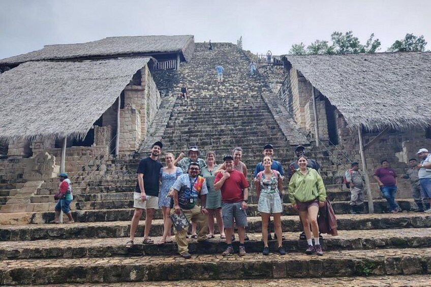 Un groupe de personnes se tient sur des marches en pierre devant une grande structure en forme de pyramide, surmontée d’un toit de chaume. 2