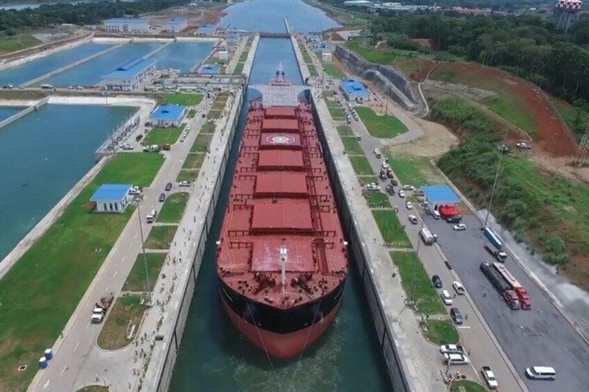 Un gran carguero navegando por un canal con esclusas, rodeado de edificios industriales y una carretera con vehículos. 4