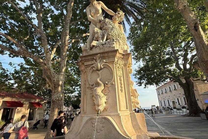 A statue with intricate carvings, a tree-lined square, and a building in the background. 3