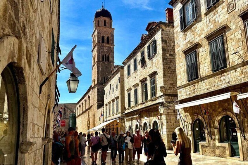 A historic street with stone buildings, a prominent tower, and pedestrians. 1