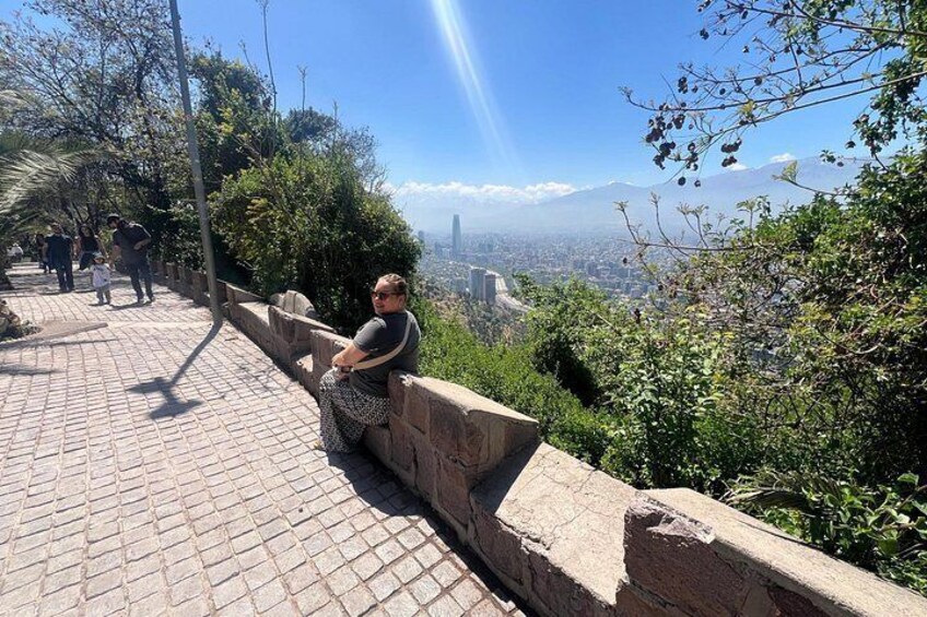 Eine Person sitzt auf einer Steinsitzbank und blickt auf eine Stadtlandschaft mit Bergen im Hintergrund. 2