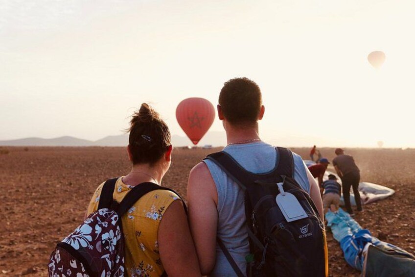 Et par sidder på stranden, med en varmluftsballon i baggrunden og mennesker i det fjerne. 3