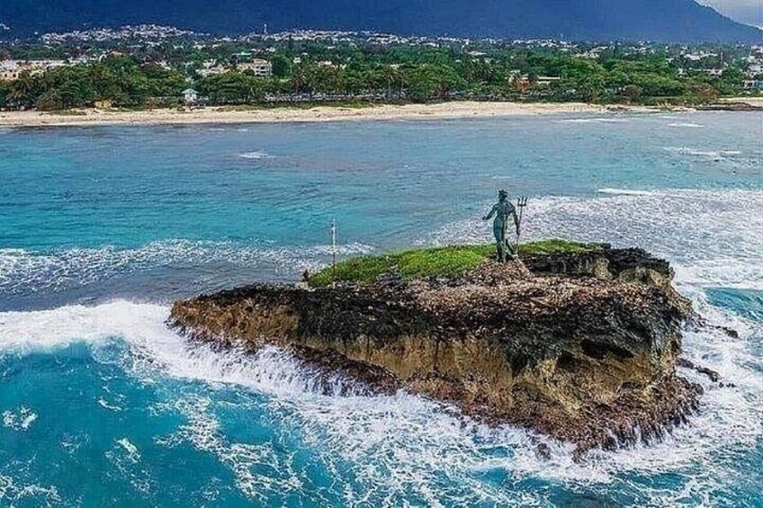 A rocky island with a statue in the ocean, surrounded by clear blue water and a distant shoreline. 4