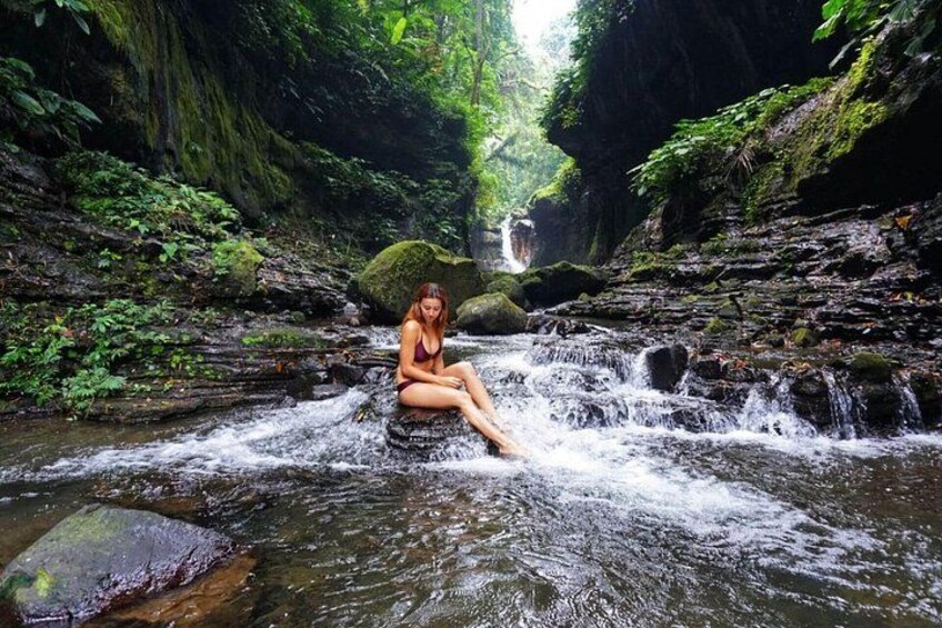 Iemand zit op een rots in een kabbelend beekje, omringd door weelderige begroeiing, met een waterval op de achtergrond. 1