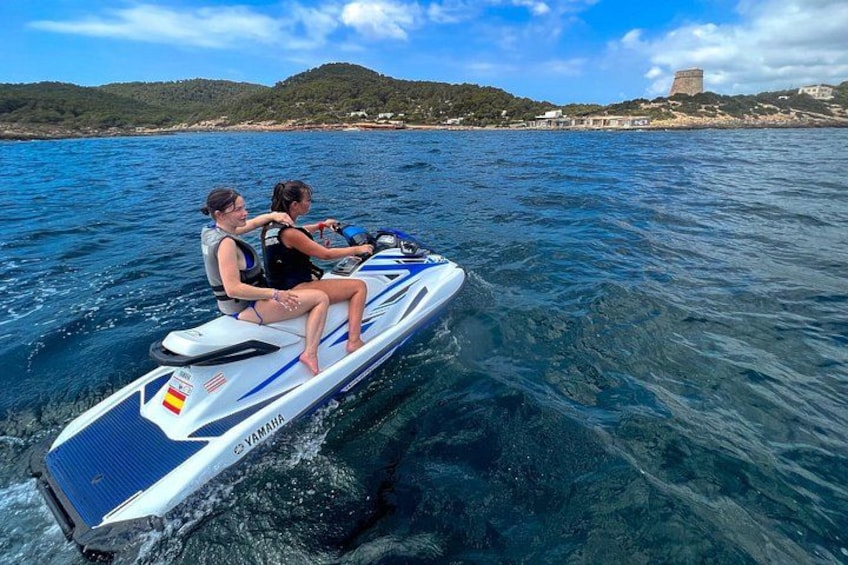 To personer på en jetski i klart vand med et bakket landskab og et tårn i baggrunden. 5