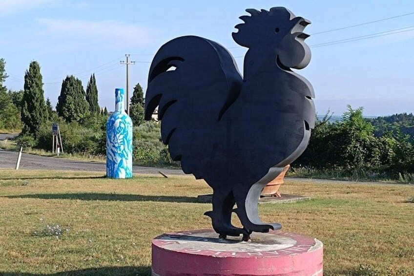 A rooster statue on a circular base, a blue and white bottle, and a road with trees in the background. 3