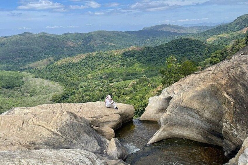 A person sitting on a rock overlooking a valley with greenery and distant hills. 1