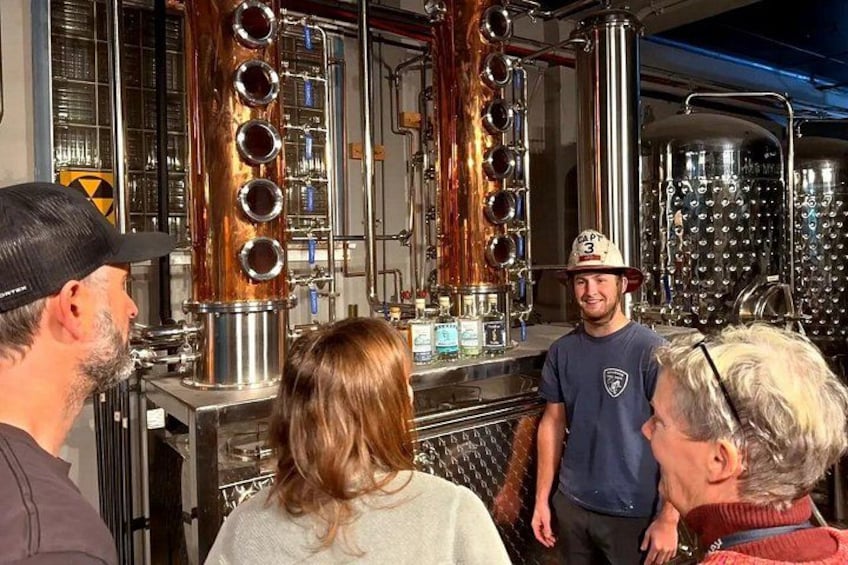 A group of people in a brewery setting with stainless steel tanks and copper pipes. 2