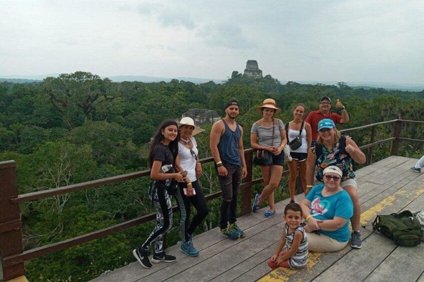 A group of people on a wooden platform overlooking a lush green landscape with a pyramid-like structure in the distance. 2