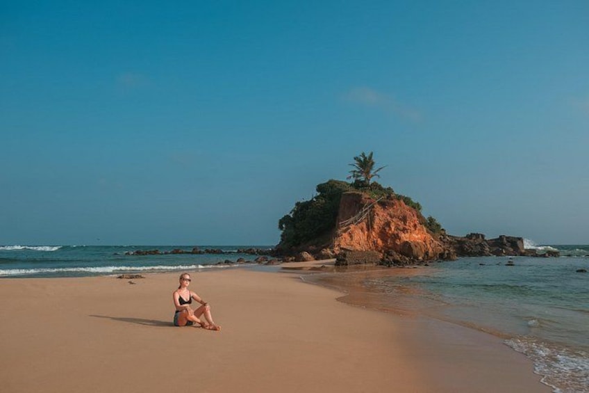 A beach with a person sitting on the sand, a large rock formation, and the ocean. 3