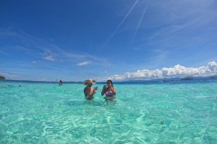 Two people wading in clear turquoise water, a clear sky, and distant boats. 3