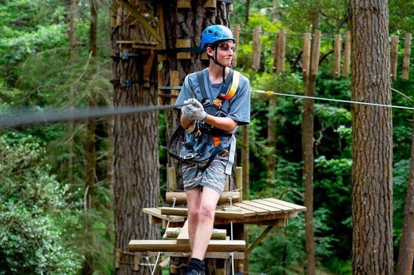 A person on a wooden platform in a forest, wearing a helmet and harness, with ropes and trees in the background. 5