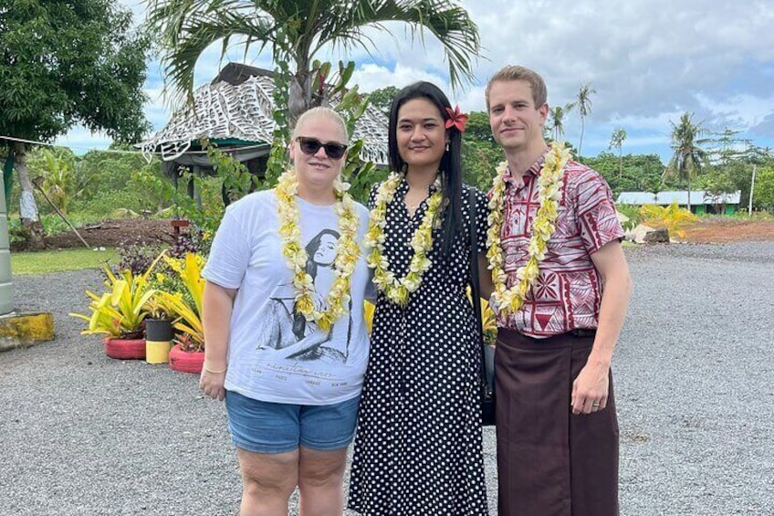Three people standing outdoors with a tropical background, including palm trees and a thatched-roof structure. 5