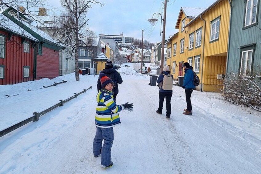 A snowy street with colorful houses, a child in a striped jacket, and two adults walking. 2