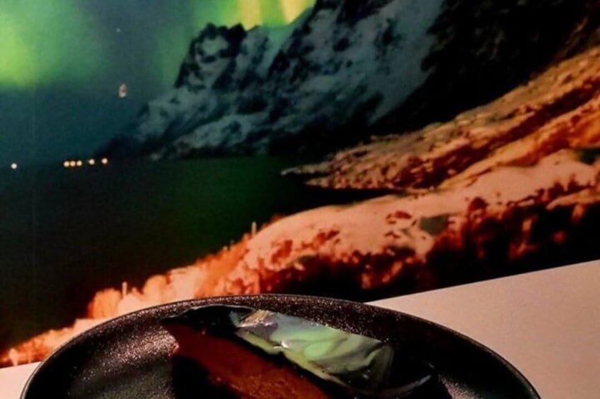 Une poêle contenant un morceau de viande et une feuille verte, le tout mis en valeur par un paysage montagneux en arrière-plan, avec un plan d’eau et des aurores boréales. 1