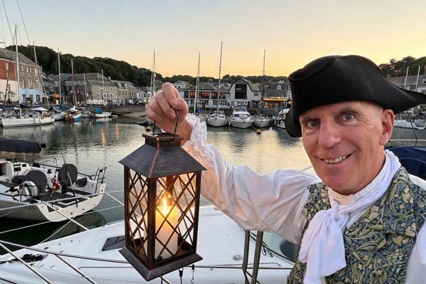 A person in historical attire holding a lantern by a marina with boats and buildings in the background. 1