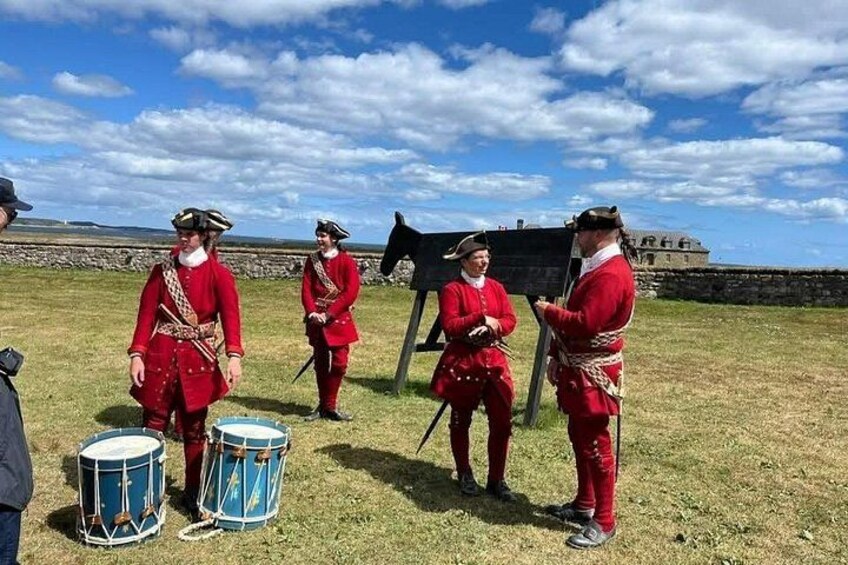 A group of people in historical uniforms standing in a grassy field with a fort in the background. 4