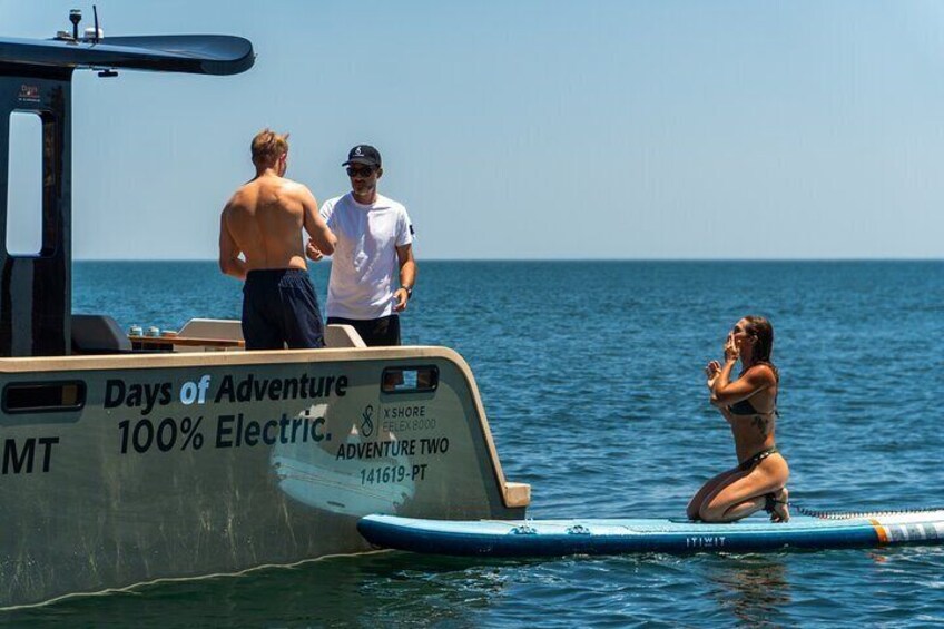 Un barco llamado 'Days of Adventure' con gente a bordo y una mujer practicando surf en el agua. 4