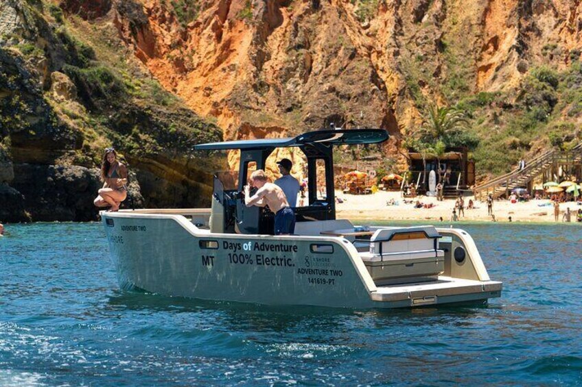 Un barco llamado 'Days of Adventure' con la leyenda '100% Electric' y 'Adventure Two' en su costado, navegando cerca de una costa rocosa con una playa al fondo. 5