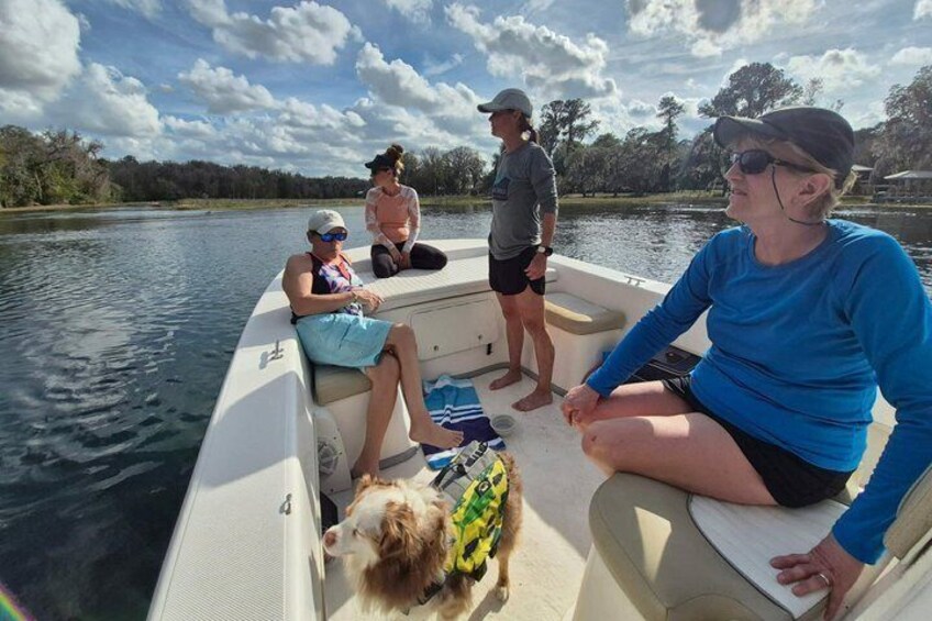 A group of people on a boat with a dog on board, surrounded by water and trees. 5