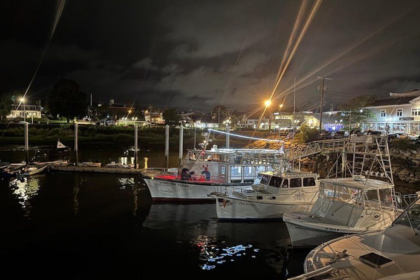Een jachthaven in de nacht met afgemeerde boten, een vuurtoren op de achtergrond en huizen langs de kust. 2