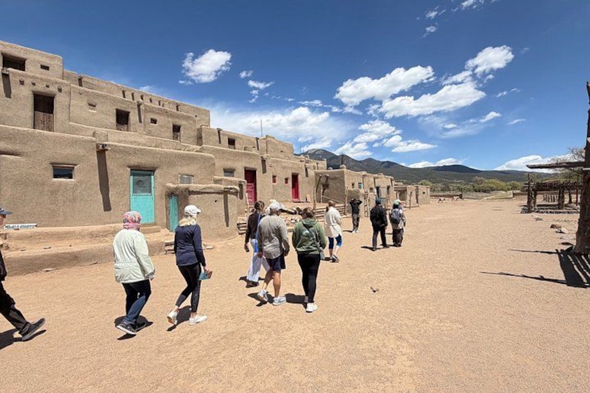 A group of people walking in a desert-like area with adobe buildings and mountains in the background. 5