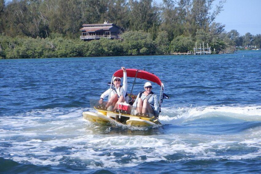 Two people on a yellow watercraft with a red canopy, moving on a body of water with trees and a house in the background. 3