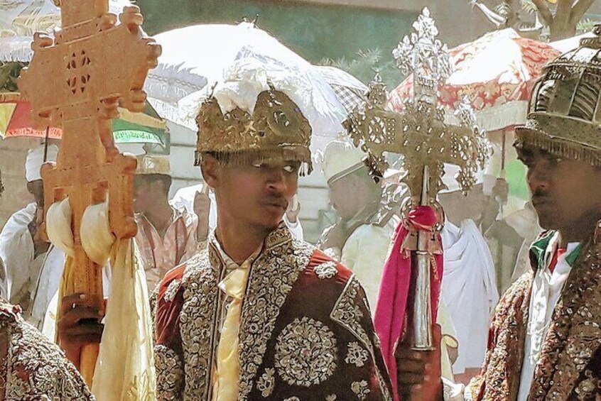 Una persona che regge un bastone elaborato e una croce cerimoniale, vestita con abiti tradizionali e decorazioni intricate, con un copricapo ornato. 2