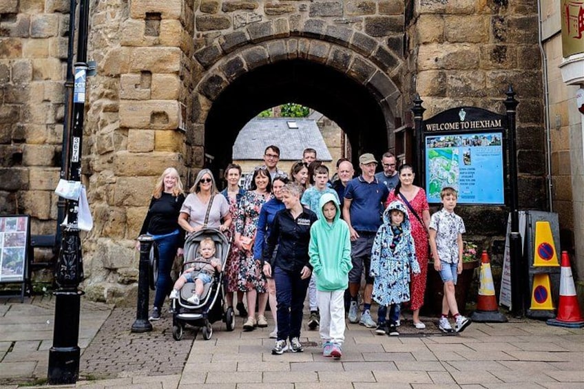 A group of people standing in front of a stone archway with a sign that reads 'WELCOME TO HEXHAM.' 3