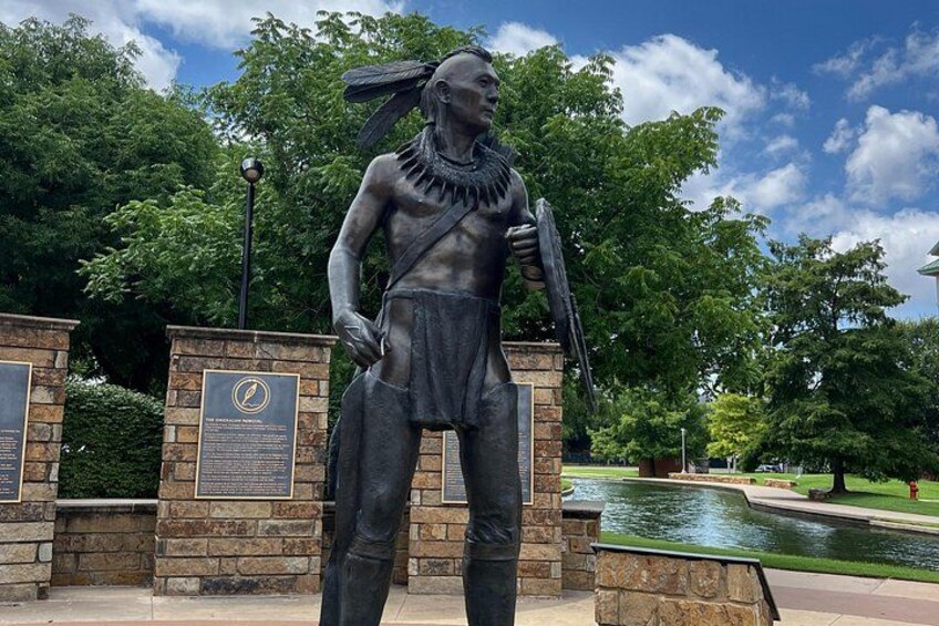 A statue of a Native American figure with a feathered headdress, standing on a stone pedestal with a plaque. 2