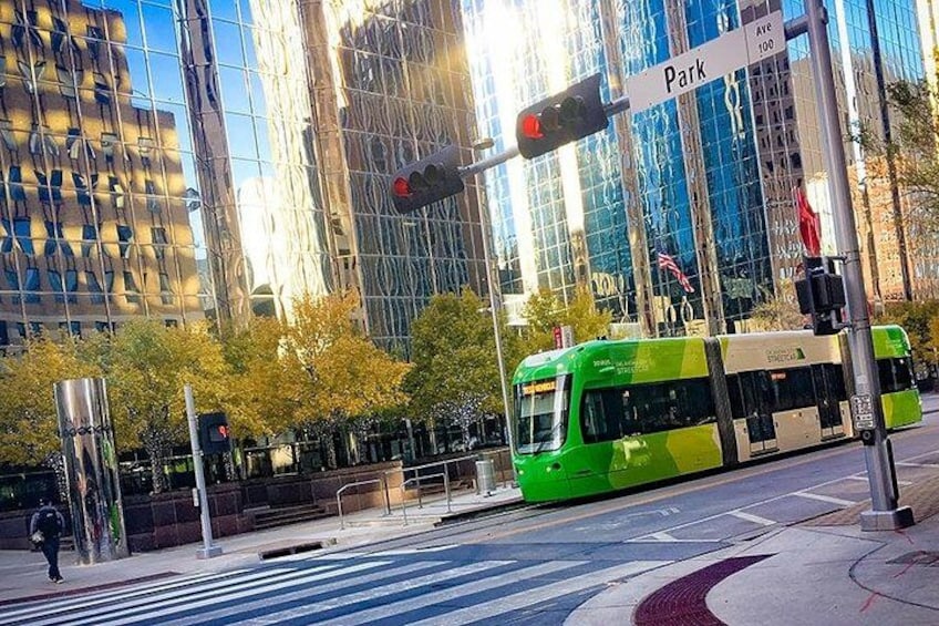 A green tram at an intersection with a traffic light, modern buildings, and trees in the background. 1