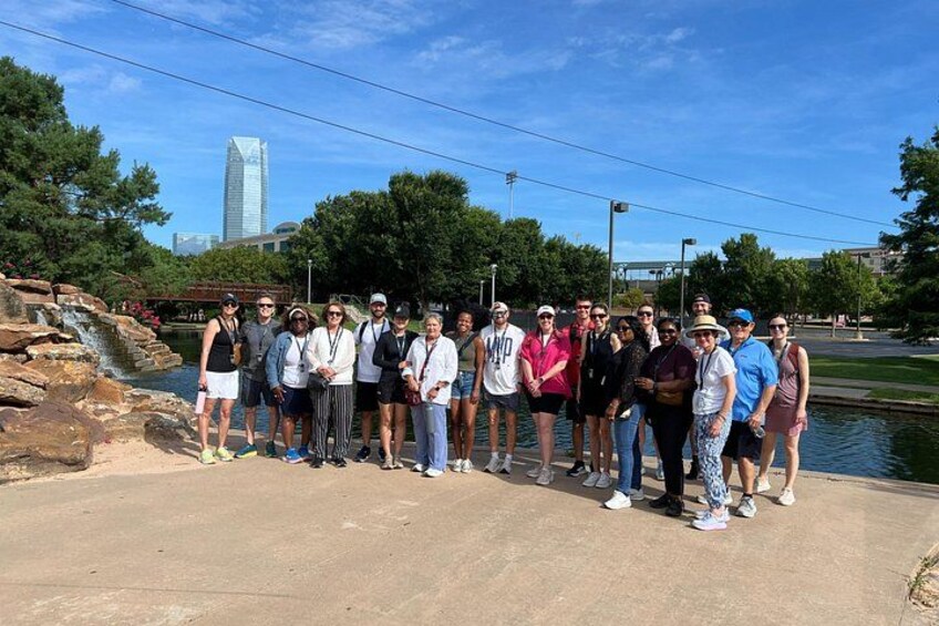 Un grupo de personas de pie sobre un sendero de concreto cerca de un cuerpo de agua, con un puente y un edificio alto al fondo. 3