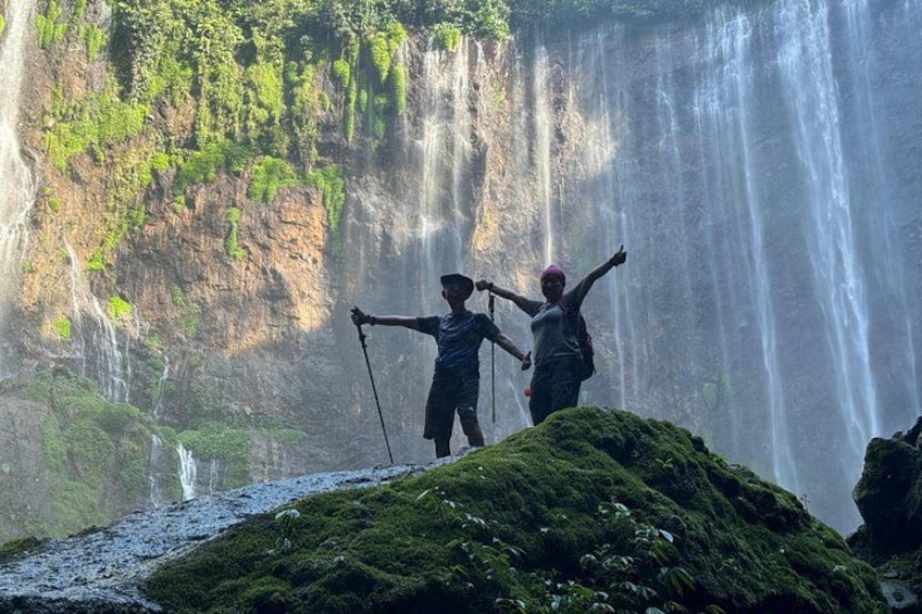 Two people standing on a moss-covered rock with a waterfall in the background. 1