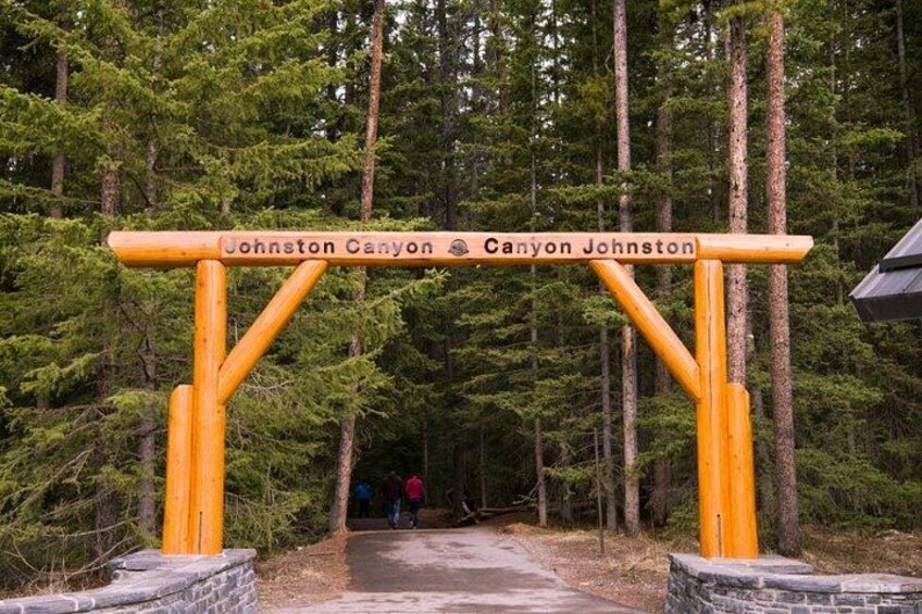 Een houten bogenpoort met 'Johnston Canyon' en 'Canyon Johnston' erop, die leidt naar een bebost gebied. 2