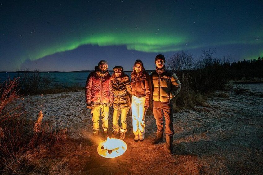 Four people standing on a beach with a campfire and the Northern Lights in the background. 4