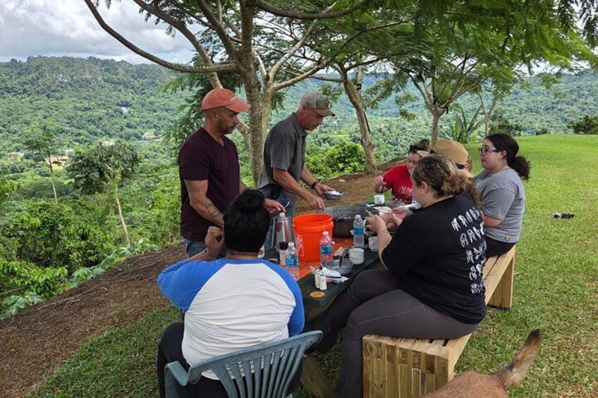 A group of people sitting at an outdoor table, engaged in conversation, with a scenic landscape in the background. 3